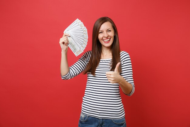 Retrato de mujer joven alegre mostrando el pulgar hacia arriba y sosteniendo un montón de dólares, dinero en efectivo aislado sobre fondo rojo brillante de la pared. Personas sinceras emociones, concepto de estilo de vida. Simulacros de espacio de copia.