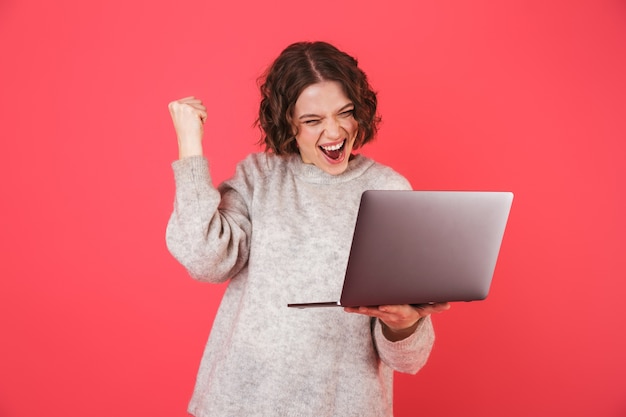 Retrato de una mujer joven alegre que se encuentran aisladas sobre rosa, trabajando en una computadora portátil, celebrando el éxito