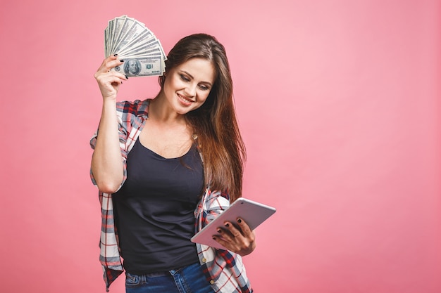Retrato de una mujer joven alegre que sostiene billetes de banco del dinero y que celebra aislado sobre fondo rosado. Usando tableta.