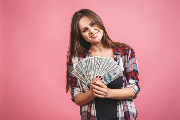 Retrato de una mujer joven alegre que sostiene billetes de banco del dinero y que celebra aislado sobre fondo rosado.
