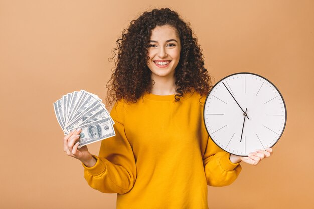 Retrato de una mujer joven alegre que sostiene billetes de banco y el reloj del dinero, celebrando aislado sobre fondo beige.