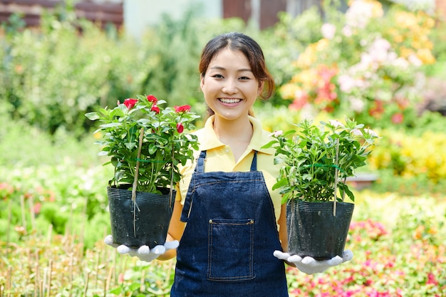 Retrato de mujer joven alegre que trabaja en el vivero de plantas, ella está sosteniendo dos macetas con flores y sonriendo a la cámara