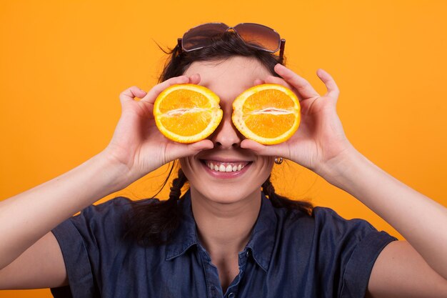 Retrato de mujer joven alegre con rodajas de naranja como ojos en estudio sobre fondo amarillo. Mujer joven bastante caucásica sosteniendo una naranja en rodajas.