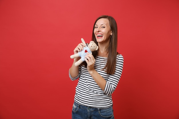 Retrato de mujer joven alegre en ropa a rayas sosteniendo y jugando con osito de peluche de juguete