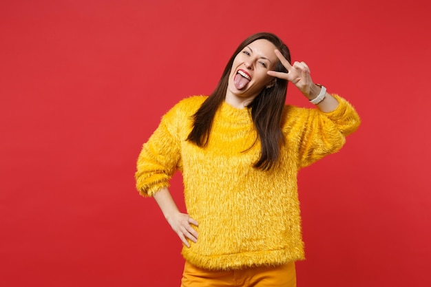 Retrato de mujer joven alegre en suéter de piel amarilla que muestra la lengua, signo de victoria aislado sobre fondo de pared rojo brillante en estudio. Personas sinceras emociones, concepto de estilo de vida. Simulacros de espacio de copia.