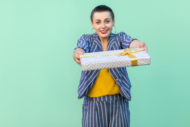 Retrato de una mujer joven amable y hermosa con el pelo corto con traje a rayas de pie, dándote, felicitándote con la caja de regalo, mirando a la cámara. Interior, aislado, foto de estudio, fondo verde