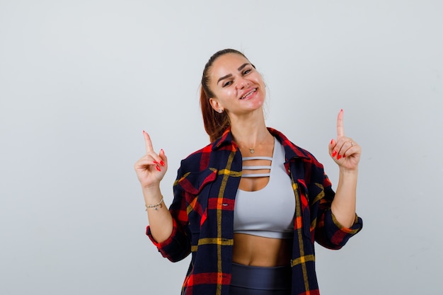 Retrato de mujer joven apuntando hacia arriba en la parte superior de la cosecha, camisa a cuadros, pantalones y mirando feliz vista frontal