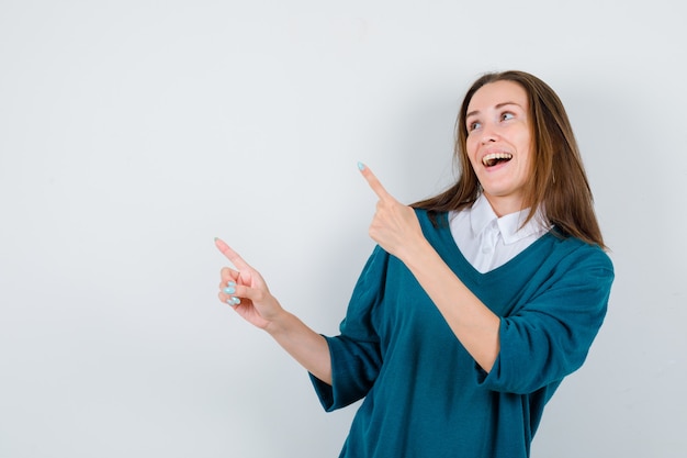 Retrato de mujer joven apuntando a la esquina superior izquierda en suéter sobre camisa y mirando feliz vista frontal