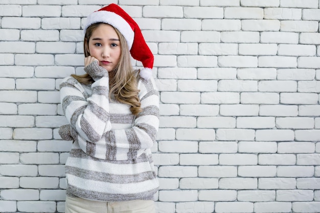 Retrato de mujer joven asiática linda en traje de Navidad y usar sombrero rojo de santa claus con sonrisa sobre fondo de ladrillo blanco y espacio de copia. Feliz de mujer adolescente en tema de Navidad.