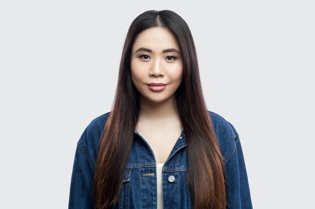 Retrato de mujer joven asiática morena hermosa en chaqueta de mezclilla azul casual con maquillaje de pie, sonriendo y mirando a cámara. tiro del estudio de interior, aislado en fondo gris claro.