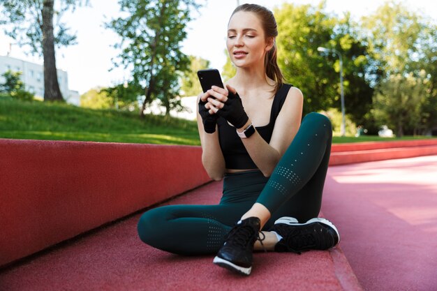 Retrato de mujer joven atlética vistiendo chándal sosteniendo el teléfono inteligente mientras está sentado en el campo de deportes durante el entrenamiento en el parque verde