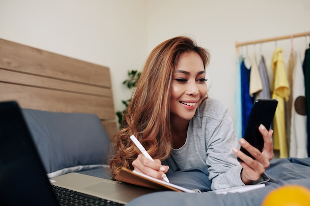 Foto retrato de mujer joven atractiva alegre comprobar mensajes de texto en el teléfono inteligente y escribir en el planificador cuando está acostado en la cómoda cama