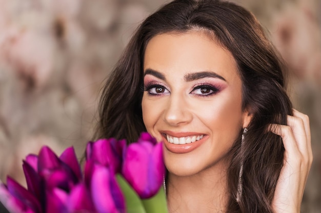 Un retrato de una mujer joven atractiva con hermosos ojos, que sostiene un ramo de tulipanes morados y representa un bonito maquillaje en su rostro.