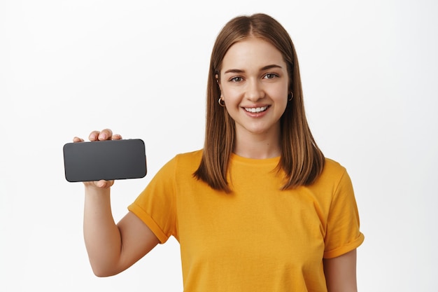 El retrato de una mujer joven atractiva muestra la pantalla horizontal del teléfono inteligente, el videojuego o la interfaz de la aplicación en el teléfono móvil, sonriendo y recomendando, pared blanca