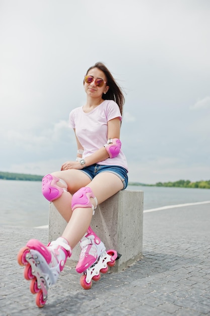 Retrato de una mujer joven y atractiva con pantalones cortos, camiseta, gafas de sol y patines sentados en el banco de hormigón en la pista de patinaje al aire libre