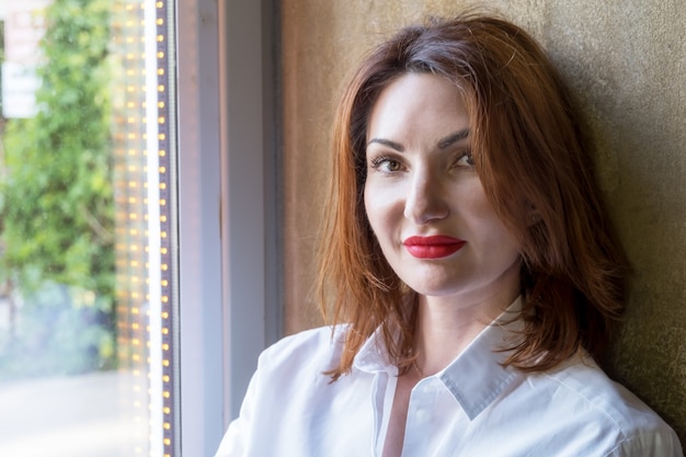 Retrato de una mujer joven atractiva con el pelo rojo con camisa blanca en el alféizar de la ventana junto a la ventana grande en un café. La mujer mira a la cámara