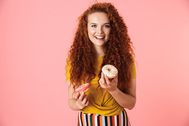 Retrato de una mujer joven atractiva con el pelo rojo largo y rizado que se encuentran aisladas, comiendo donas