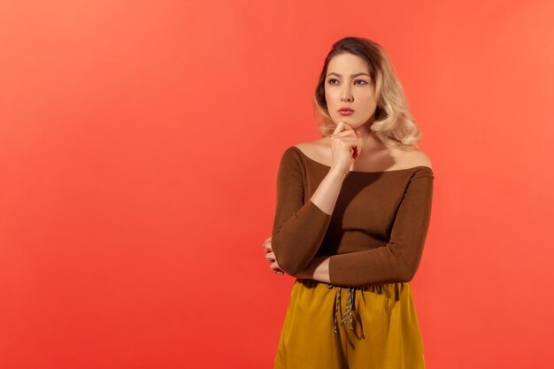 Retrato de una mujer joven en blusa marrón reflexionando con una expresión seria Ella se toca la barbilla y piensa con la cara pensativa mirando hacia otro lado Disparo de estudio interior aislado sobre fondo rojo