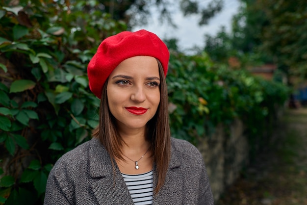 Retrato de una mujer joven con boina roja cerca de una valla de piedra cubierta de vegetación