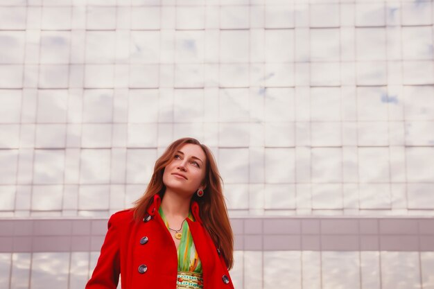 Retrato de mujer joven y bonita de apariencia eslava con vestido amarillo y abrigo rojo. Fachada del edificio comercial