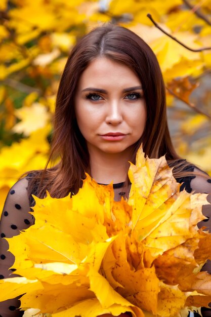 Retrato de mujer joven y bonita de apariencia eslava en vestido oscuro en otoño, de pie contra el fondo de un parque otoñal con hojas en sus manos. Linda modelo camina en el parque en otoño dorado