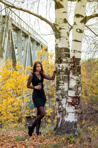 Retrato de mujer joven y bonita de apariencia eslava en vestido oscuro y vaso de café en otoño, de pie junto al árbol de abedul contra el fondo