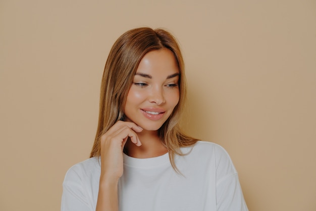 Retrato de mujer joven y bonita con cabello castaño claro recto en camiseta blanca casual de manga larga, manteniendo una mano en el pómulo mirando el espacio en blanco con una sonrisa, aislado sobre fondo beige pastel