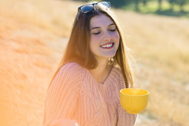 Retrato de una mujer joven y bonita con un elegante suéter de punto de pie en el bosque dorado de otoño en un día soleado Tiempo para relajarse y tomar un café