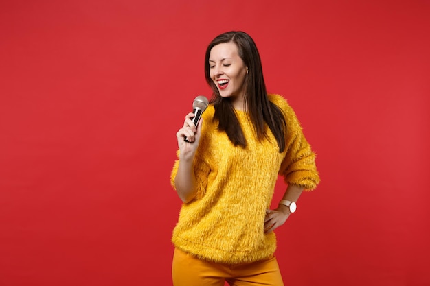 Retrato de mujer joven y bonita impresionante en suéter de piel amarilla canta la canción en el micrófono aislado sobre fondo de pared rojo brillante en estudio. Personas sinceras emociones, concepto de estilo de vida. Simulacros de espacio de copia.