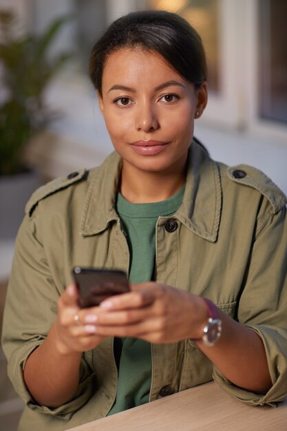 Retrato de mujer joven y bonita mirando a la cámara mientras usa el teléfono móvil en la mesa