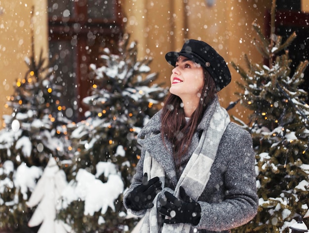 Retrato de una mujer joven y bonita que usa un elegante sombrero negro antes del fondo de la ciudad de Navidad en tiempo de nieve