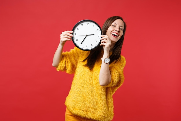 Retrato de mujer joven y bonita de risa en suéter de piel amarillo con reloj redondo aislado sobre fondo de pared rojo brillante en estudio. Personas sinceras emociones, concepto de estilo de vida. Simulacros de espacio de copia.