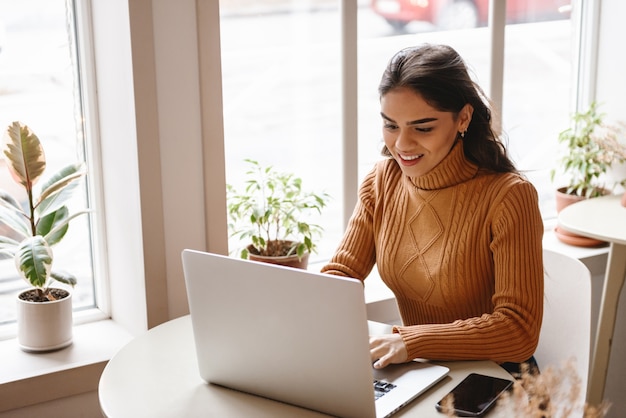 Retrato de una mujer joven y bonita sentada en un café en el interior con ordenador portátil.