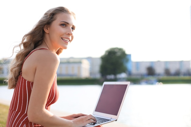 Retrato de mujer joven y bonita sentada en la orilla del río durante el día de verano, usando la computadora portátil.