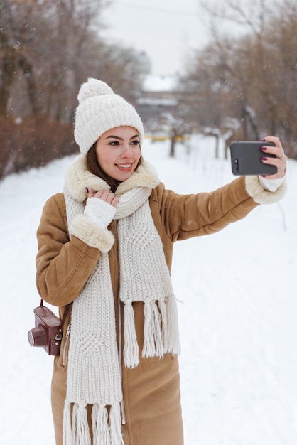 Retrato de una mujer joven y bonita con sombrero y bufanda caminando al aire libre en la nieve del invierno con teléfono móvil tomar un selfie.