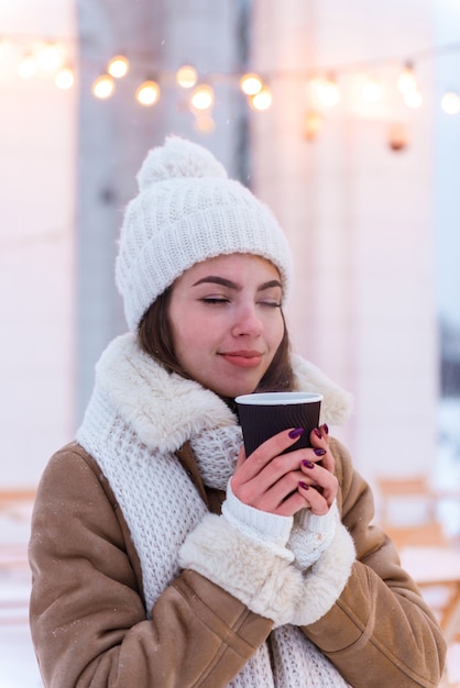 Retrato de una mujer joven y bonita con sombrero y bufanda caminando al aire libre en la nieve del invierno tomando café.