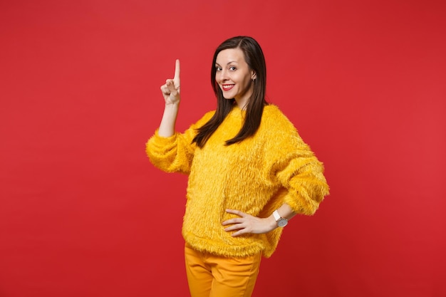 Retrato de mujer joven y bonita sonriente en suéter de piel amarilla apuntando con el dedo índice hacia arriba aislado sobre fondo rojo brillante de la pared en estudio. Personas sinceras emociones, concepto de estilo de vida. Simulacros de espacio de copia.