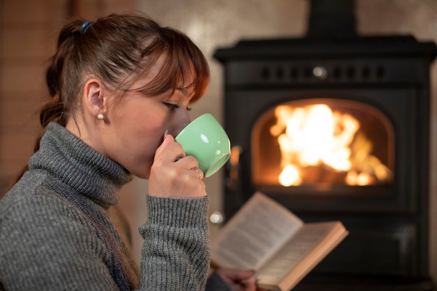 Foto retrato de mujer joven y bonita tomando café y leyendo un libro cerca de la chimenea