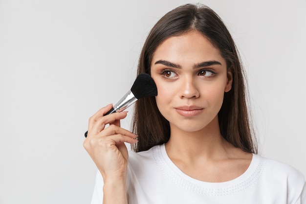 Retrato de una mujer joven y bonita vestida casualy aislado en blanco, aplicando maquillaje con un pincel