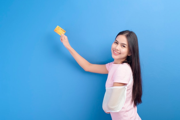 Un retrato de una mujer joven con un brazo lesionado en un cabestrillo sosteniendo una tarjeta de crédito o tarjeta de seguro médico sobre fondo azul en el estudio, el seguro y el concepto de salud