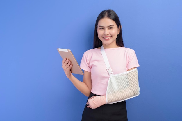 Un retrato de una mujer joven con un brazo lesionado en un cabestrillo usando una tableta sobre fondo azul en el concepto de estudio, seguro y atención médica