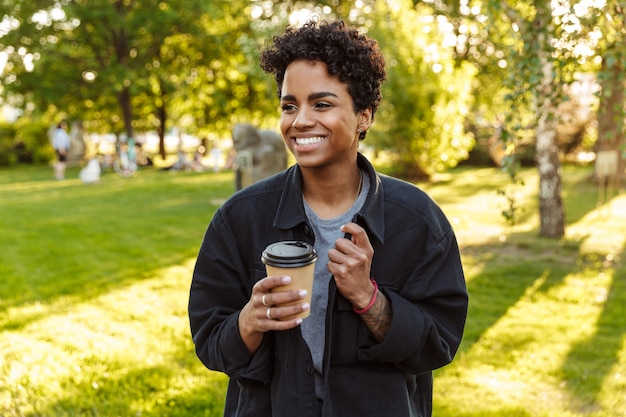 Retrato de mujer joven con cabello rizado sosteniendo y bebiendo café para llevar de un vaso de papel mientras camina en el parque de la ciudad