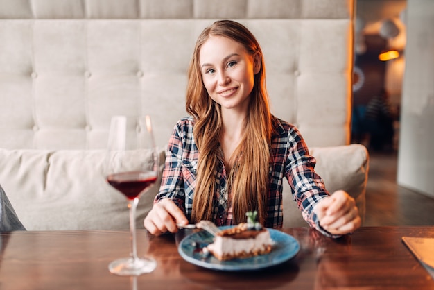 Retrato de mujer joven en café, pastel dulce y vino tinto en un vaso sobre la mesa. Chica con postre de chocolate en restaurante.