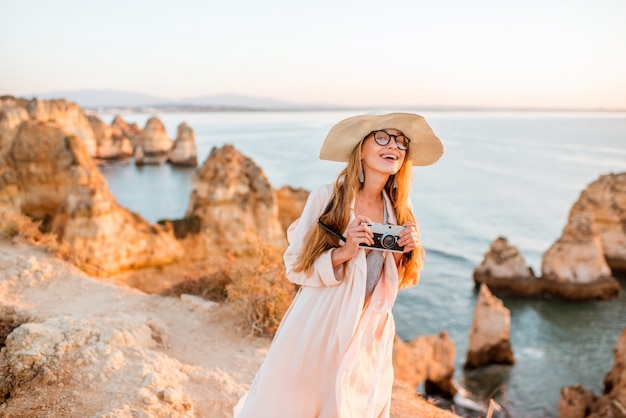 Retrato de una mujer joven con cámara de fotos disfrutando de una gran vista de la costa rocosa durante el amanecer en Lagos, en el sur de Portugal