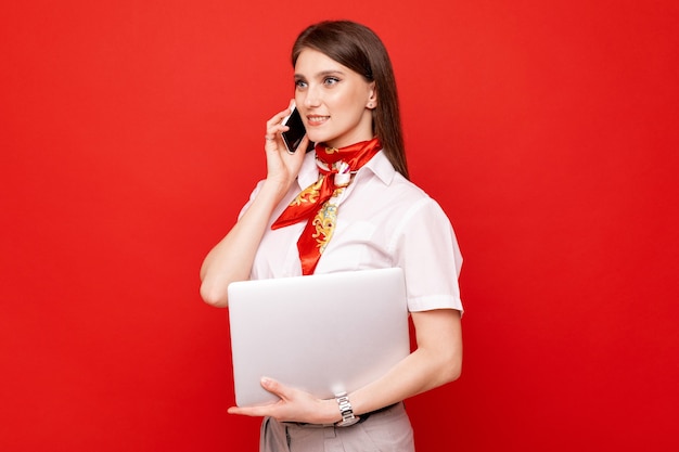 Retrato de una mujer joven con una camisa blanca sosteniendo una computadora portátil y hablando por teléfono en una pared roja