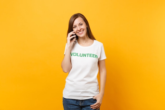 Retrato de mujer joven en camiseta blanca con inscripción escrita voluntario de título verde hablando por teléfono móvil aislado sobre fondo amarillo. Ayuda de asistencia gratuita voluntaria, concepto de trabajo de gracia de caridad.