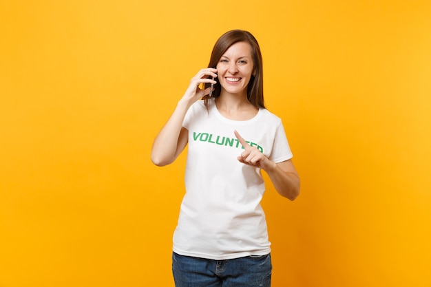 Retrato de mujer joven en camiseta blanca con inscripción escrita voluntario de título verde hablando por teléfono móvil aislado sobre fondo amarillo. Ayuda de asistencia gratuita voluntaria, concepto de trabajo de gracia de caridad.