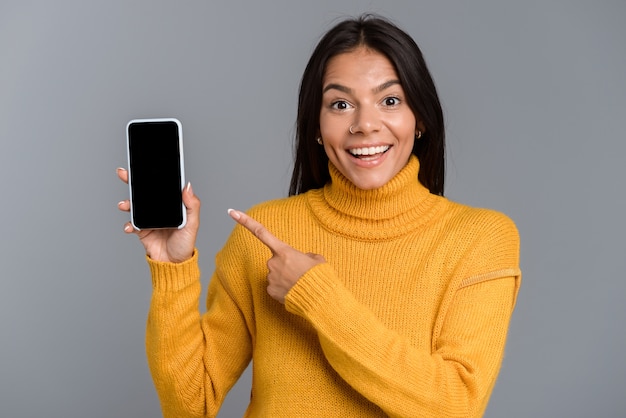 Retrato de una mujer joven casual feliz aislada sobre pared gris, mostrando teléfono móvil de pantalla en blanco