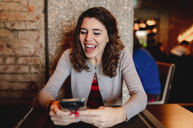 Retrato de mujer joven caucásica sonriente en un restaurante sosteniendo y usando un teléfono inteligente.