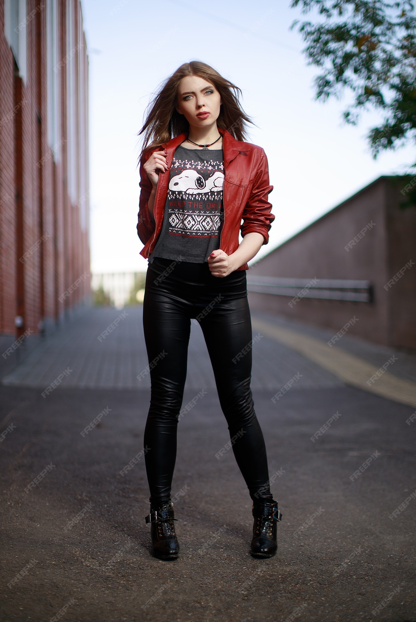 El retrato de una mujer joven con una chaqueta de cuero roja leggings negros permanece en la ciudad durante el día. | Foto Premium